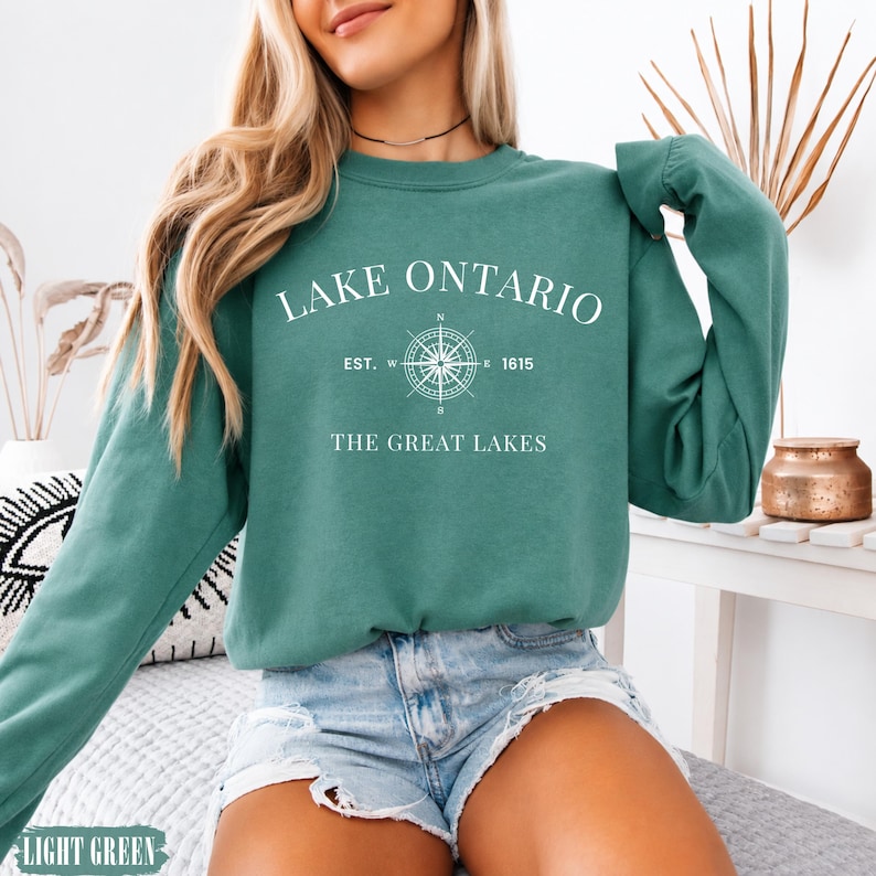 May include: A teal long-sleeve crewneck sweatshirt featuring white text that reads LAKE ONTARIO, EST. 1615, and THE GREAT LAKES, centred around a white compass graphic. The sweatshirt has a ribbed neckline, cuffs, and hem, with a solid colour finish.