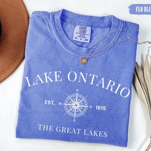May include: A periwinkle blue short-sleeved t-shirt with white text reading LAKE ONTARIO, EST. 1615, and THE GREAT LAKES, featuring a central white compass rose graphic. The shirt has a crew neckline and a Comfort Colors brand label at the neck.