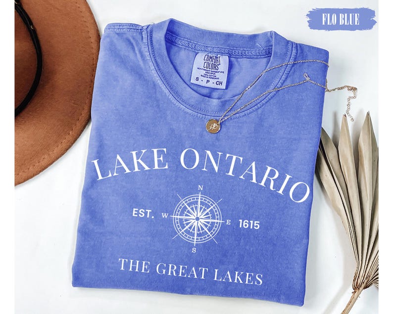 May include: A periwinkle blue short-sleeved t-shirt with white text reading LAKE ONTARIO, EST. 1615, and THE GREAT LAKES, featuring a central white compass rose graphic. The shirt has a crew neckline and a Comfort Colors brand label at the neck.