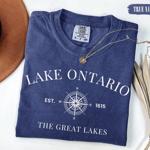 May include: A true navy short-sleeved t-shirt featuring white text that reads LAKE ONTARIO, EST. 1615, and THE GREAT LAKES, centred around a white compass rose graphic. The shirt has a crew neckline and a Comfort Colors brand label at the collar.