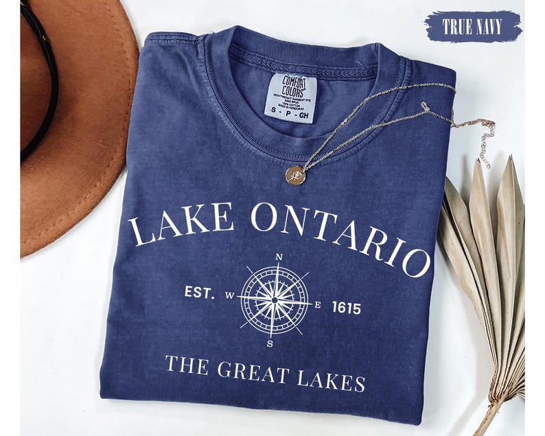 May include: A true navy short-sleeved t-shirt featuring white text that reads LAKE ONTARIO, EST. 1615, and THE GREAT LAKES, centred around a white compass rose graphic. The shirt has a crew neckline and a Comfort Colors brand label at the collar.