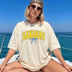 May include: Cream-colored t-shirt with the word "BANANAS" in yellow block letters outlined in blue. A blue bow graphic is centered below the word. The shirt is worn on a boat with a blue sky and water in the background.
