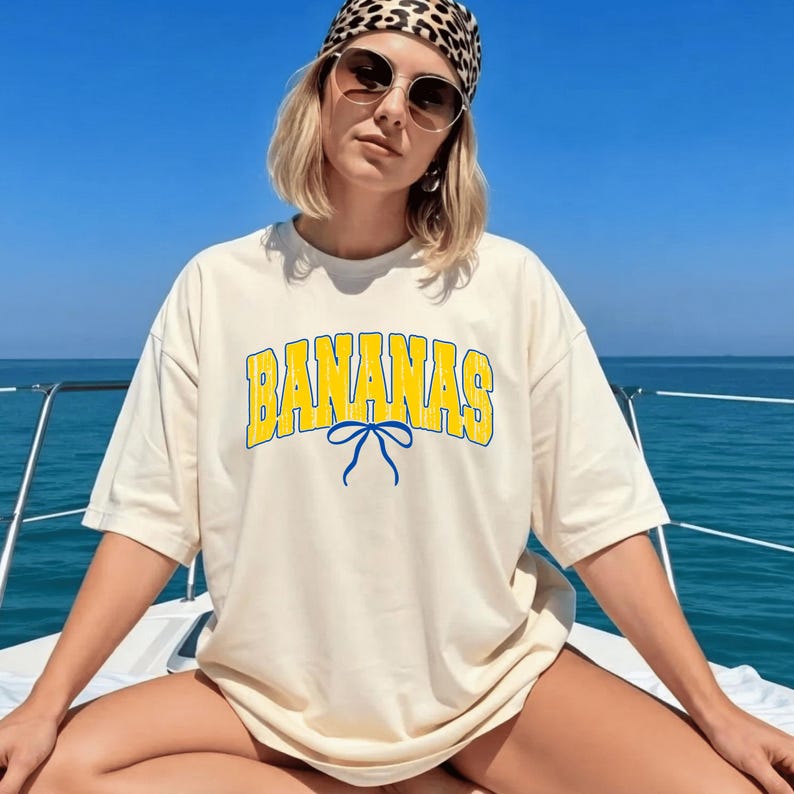 May include: Cream-colored t-shirt with the word "BANANAS" in yellow block letters outlined in blue. A blue bow graphic is centered below the word. The shirt is worn on a boat with a blue sky and water in the background.
