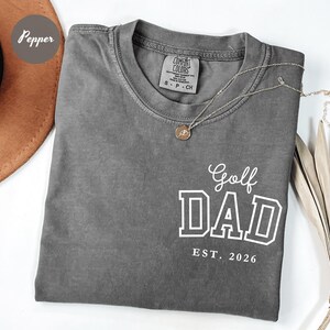 May include: Grey Comfort Colors t-shirt with the text "Golf DAD EST. 2026" in white. A gold necklace with a pendant is draped across the shirt. The shirt is folded and displayed on a white surface.