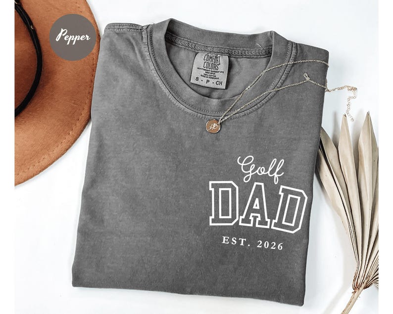 May include: Grey Comfort Colors t-shirt with the text "Golf DAD EST. 2026" in white. A gold necklace with a pendant is draped across the shirt. The shirt is folded and displayed on a white surface.