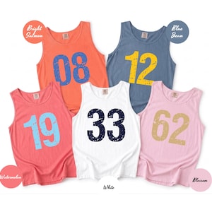 May include: Five sleeveless tank tops in various colors: bright salmon, blue jean, watermelon, white, and blossom. Each tank top features a large number in a contrasting color: 08, 12, 19, 33, and 62. The tank tops are casual wear.