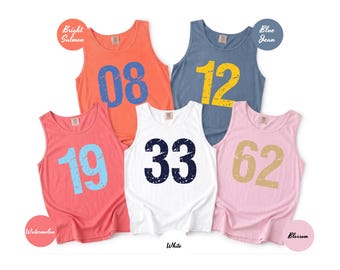 May include: Five sleeveless tank tops in various colors: bright salmon, blue jean, watermelon, white, and blossom. Each tank top features a large number in a contrasting color: 08, 12, 19, 33, and 62. The tank tops are casual wear.