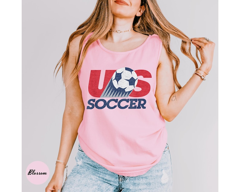 May include: Pink tank top with the text "US SOCCER" in red and navy blue. The design includes a soccer ball with blue and white accents. The tank top is sleeveless and made of a soft material.