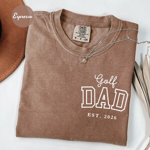 May include: A brown t-shirt with the text "Golf DAD EST. 2026" in white. The shirt has a crew neck and short sleeves. A silver necklace with a small pendant is draped across the shirt. The shirt is a shade of brown.