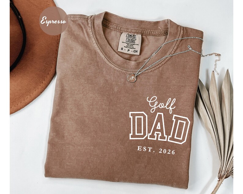 May include: A brown t-shirt with the text "Golf DAD EST. 2026" in white. The shirt has a crew neck and short sleeves. A silver necklace with a small pendant is draped across the shirt. The shirt is a shade of brown.