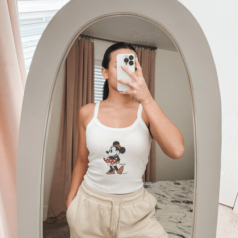 May include: A white ribbed tank top featuring a graphic print of Minnie Mouse wearing a red polka dot skirt and brown shoes. The tank top has a scoop neckline and thick shoulder straps, paired with beige drawstring sweatpants.