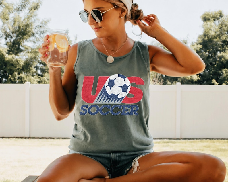 May include: A gray tank top with the words "US SOCCER" in red and blue, featuring a soccer ball graphic. The tank top is worn by a person wearing sunglasses.
