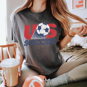 Peut inclure: T-shirt gris fonc&eacute; avec l'inscription "US SOCCER" en rouge et bleu, avec un graphisme de ballon de football. Le t-shirt est port&eacute; avec un legging gris et des chaussettes orange. Une tasse en plastique transparent avec une paille et une boisson est au premier plan.