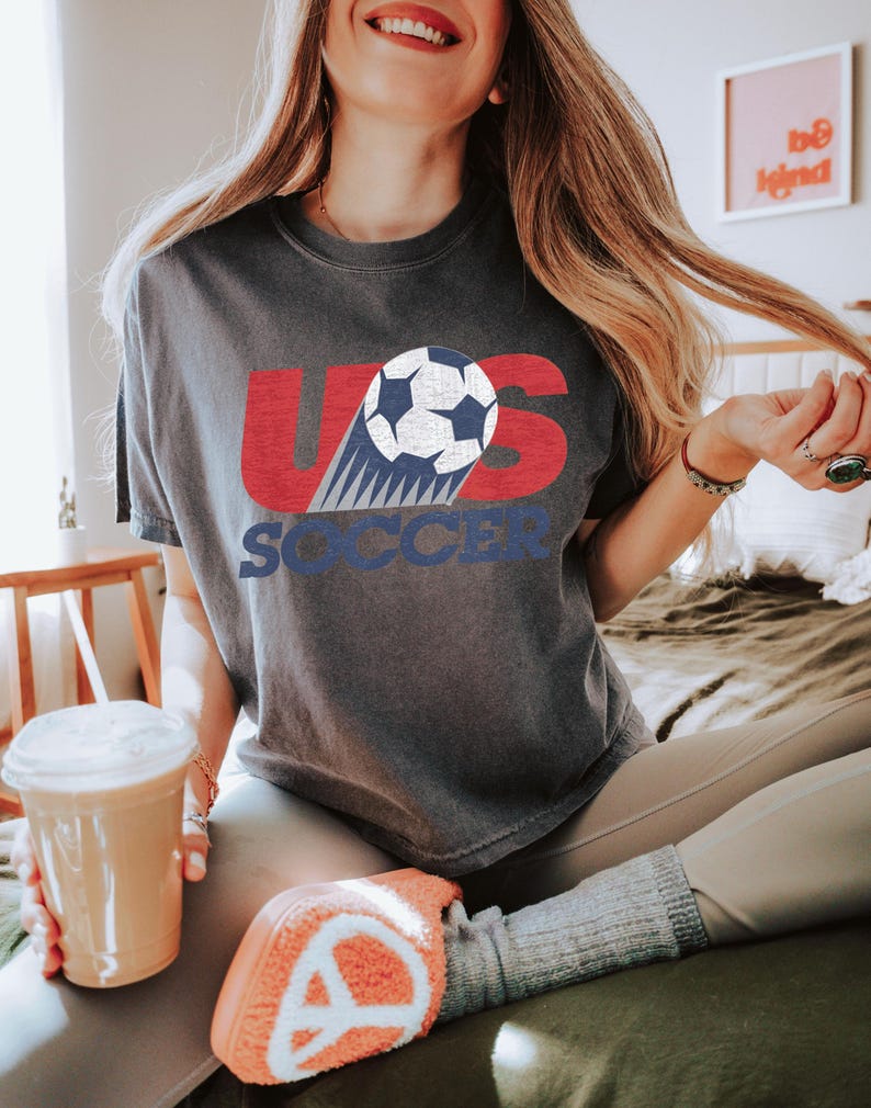 Peut inclure: T-shirt gris fonc&eacute; avec l'inscription "US SOCCER" en rouge et bleu, avec un graphisme de ballon de football. Le t-shirt est port&eacute; avec un legging gris et des chaussettes orange. Une tasse en plastique transparent avec une paille et une boisson est au premier plan.