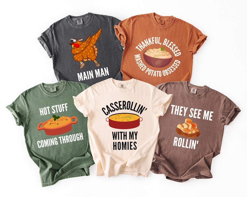 May include: Five short-sleeve t-shirts in charcoal, orange, olive green, cream, and brown. Graphics include a dabbing turkey with 'MAIN MAN', mashed potatoes with 'THANKFUL, BLESSED, MASHED POTATO OBSESSED', a casserole with 'HOT STUFF COMING THROUGH', 'CASSEROLLIN' WITH MY HOMIES', and rolls with 'THEY SEE ME ROLLIN'.