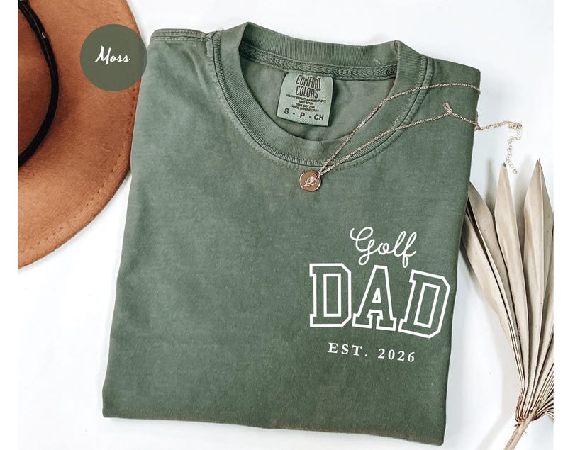 May include: A moss green t-shirt with the words "Golf DAD EST. 2026" in white block letters. A gold necklace with a circular pendant rests on the shirt. The shirt is folded on a white surface.