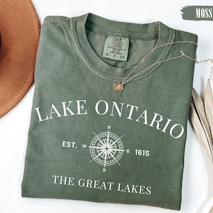 May include: A moss green short-sleeve t-shirt featuring white text that reads LAKE ONTARIO, EST. 1615, and THE GREAT LAKES, along with a white compass graphic. The shirt has a crew neckline and a Comfort Colors brand label inside the collar.