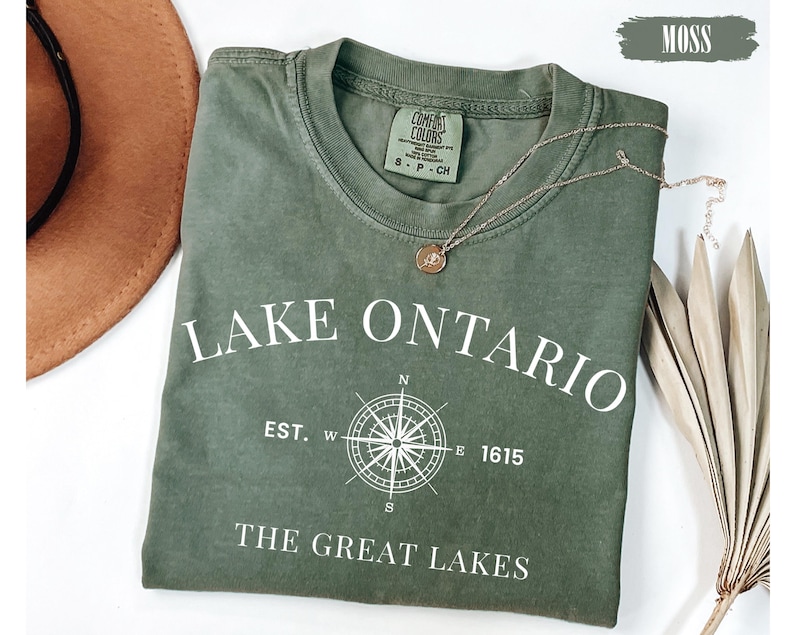 May include: A moss green short-sleeve t-shirt featuring white text that reads LAKE ONTARIO, EST. 1615, and THE GREAT LAKES, along with a white compass graphic. The shirt has a crew neckline and a Comfort Colors brand label inside the collar.