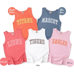 May include: Five sleeveless tank tops in various colors: bright salmon, blue jean, watermelon, white, and blossom. Each tank top features a different team name in a collegiate font: TITANS, MASCOT, LIONS, TIGERS, and EAGLES. The tank tops are displayed on a white background.
