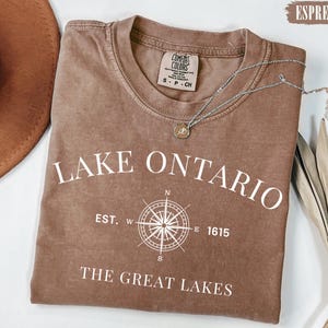 May include: An espresso-coloured short-sleeve t-shirt featuring white screen-printed text that reads LAKE ONTARIO, EST. 1615, and THE GREAT LAKES, along with a central compass rose graphic. The shirt has a crew neckline and a Comfort Colors brand label.
