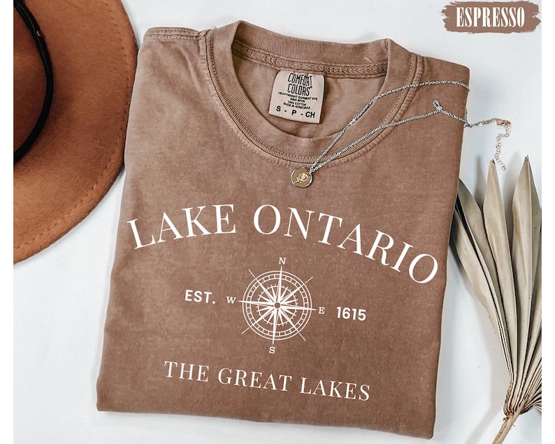 May include: An espresso-coloured short-sleeve t-shirt featuring white screen-printed text that reads LAKE ONTARIO, EST. 1615, and THE GREAT LAKES, along with a central compass rose graphic. The shirt has a crew neckline and a Comfort Colors brand label.