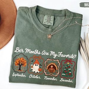 May include: A moss green crewneck t-shirt featuring the text "Ber Months Are My Favourite" above four framed illustrations representing September, October, November, and December with seasonal icons including a tree, ghost, turkey, and Christmas tree.