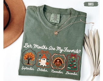 May include: A moss green crewneck t-shirt featuring the text "Ber Months Are My Favorite" above four framed illustrations representing September, October, November, and December with seasonal icons including a tree, ghost, turkey, and Christmas tree.