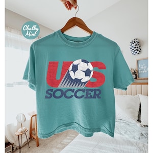 Peut inclure: Un t-shirt turquoise avec l'inscription "US SOCCER" en rouge et bleu. Le motif repr&eacute;sente un ballon de football avec une tra&icirc;n&eacute;e bleue et blanche. Le t-shirt est suspendu &agrave; un cintre en bois.