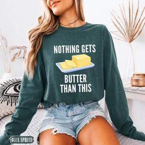 May include: A dark green long-sleeve sweatshirt featuring a graphic of a yellow stick of butter on a white plate. The text above the graphic reads "NOTHING GETS" and the text below reads "BUTTER THAN THIS" in white capital letters.