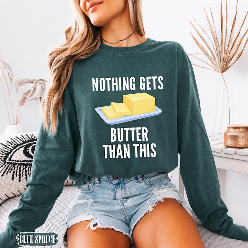May include: A dark green long-sleeve sweatshirt featuring a graphic of a yellow stick of butter on a white plate. The text above the graphic reads "NOTHING GETS" and the text below reads "BUTTER THAN THIS" in white capital letters.