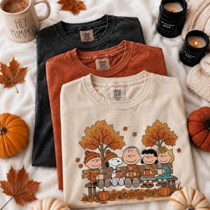 May include: Three crewneck t-shirts in black, rust orange, and cream. The cream shirt features a graphic of cartoon characters sitting on a bench under autumn trees, holding mugs. Each shirt has a Comfort Colors label at the inner neckline.