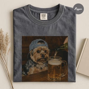 May include: A charcoal gray crew neck t-shirt featuring a graphic print of a small brown and black dog wearing a blue baseball cap, sitting at a wooden table next to a glass mug being filled with beer. The shirt has a Comfort Colors brand label.