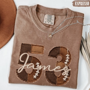 May include: A brown short-sleeve t-shirt featuring the number 53 in a patchwork football-themed design with white stitching details. The name James is written in white cursive script across the numbers. The shirt has a Comfort Colors brand label.