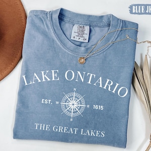 May include: A blue jean colored t-shirt featuring white text that reads LAKE ONTARIO, EST. 1615, and THE GREAT LAKES, with a central compass rose graphic. The shirt has a crew neckline and a Comfort Colors brand label on the inside collar.