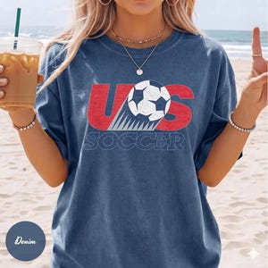 May include: Denim blue t-shirt with a graphic design featuring the text "US SOCCER" in red and white. The design includes a soccer ball. The shirt is a casual style.
