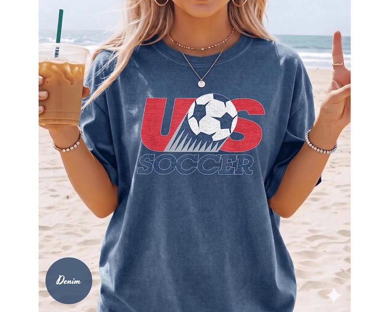 May include: Denim blue t-shirt with a graphic design featuring the text "US SOCCER" in red and white. The design includes a soccer ball. The shirt is a casual style.