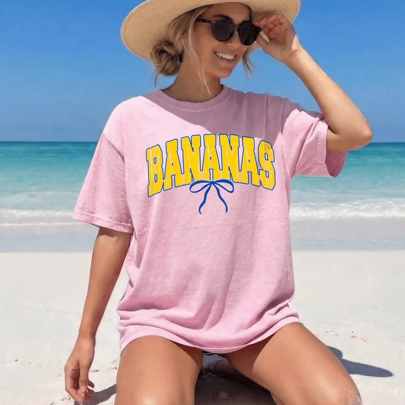 May include: A light pink t-shirt with the word "BANANAS" in yellow block letters with a blue outline and a blue bow. The shirt is worn by a person wearing sunglasses and a straw hat.