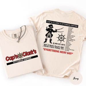 May include: An ivory t-shirt featuring a graphic of a pirate with a hook and sword, a ship wheel, and the text Cap'n Clark's Ottoman Empire. The back lists furniture items and a phone number, while the front displays the brand name in red and black.