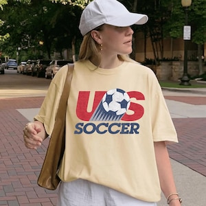 May include: A light yellow t-shirt with the text "US SOCCER" in red and blue, featuring a soccer ball graphic. The shirt is worn with a white cap and a brown shoulder bag.