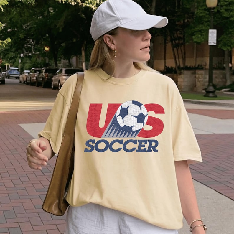 May include: A light yellow t-shirt with the text "US SOCCER" in red and blue, featuring a soccer ball graphic. The shirt is worn with a white cap and a brown shoulder bag.