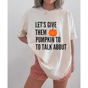 May include: An off-white short-sleeve t-shirt featuring the black text LET'S GIVE THEM PUMPKIN TO TO TALK ABOUT with an orange pumpkin graphic replacing the word pumpkin. The shirt has a crew neckline and a loose, relaxed fit throughout the torso.