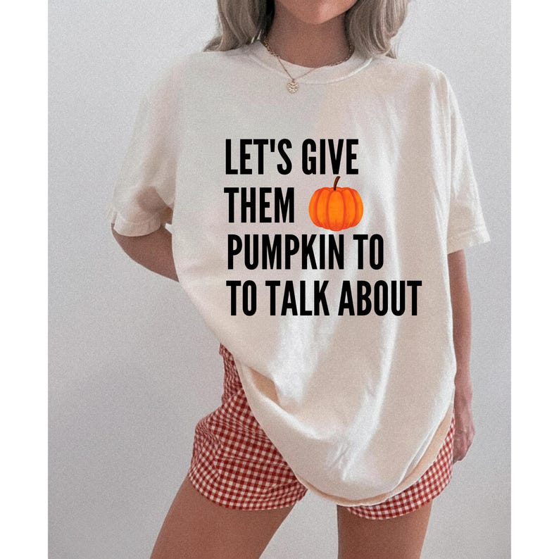 May include: An off-white short-sleeve t-shirt featuring the black text LET'S GIVE THEM PUMPKIN TO TO TALK ABOUT with an orange pumpkin graphic replacing the word pumpkin. The shirt has a crew neckline and a loose, relaxed fit throughout the torso.
