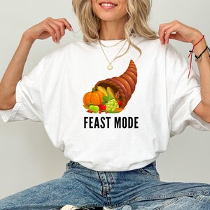 May include: A white short-sleeve t-shirt featuring a graphic of a brown cornucopia overflowing with orange pumpkins, yellow corn, green grapes, and red berries. Below the cornucopia, the text FEAST MODE is printed in bold black capital letters.