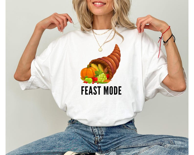 May include: A white short-sleeve t-shirt featuring a graphic of a brown cornucopia overflowing with orange pumpkins, yellow corn, green grapes, and red berries. Below the cornucopia, the text FEAST MODE is printed in bold black capital letters.