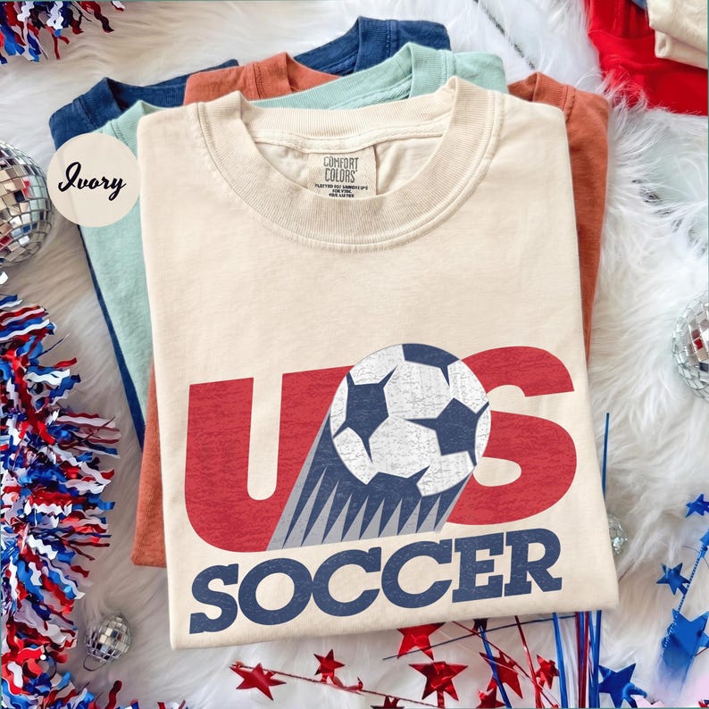 May include: Ivory-colored t-shirt with "US SOCCER" in red and blue, featuring a soccer ball graphic. The shirt is part of a stack of folded t-shirts in various colors, including blue, green, and orange. The shirts are on a white, fluffy surface.