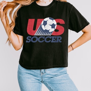 Peut inclure: T-shirt noir avec un graphisme repr&eacute;sentant un ballon de football et le texte "US SOCCER" en rouge, blanc et bleu. Le t-shirt a des manches courtes et un col rond.