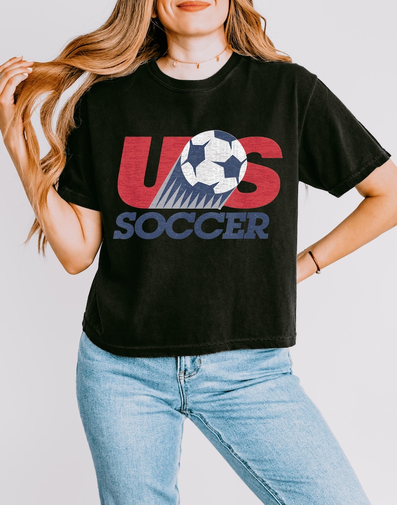 Peut inclure: T-shirt noir avec un graphisme repr&eacute;sentant un ballon de football et le texte "US SOCCER" en rouge, blanc et bleu. Le t-shirt a des manches courtes et un col rond.