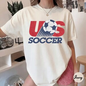 May include: Ivory-colored t-shirt with the text "US SOCCER" in red and blue. The design includes a soccer ball and a trail of blue and white lines, creating a dynamic visual.