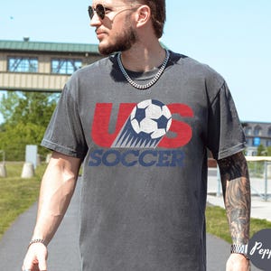 May include: A charcoal gray t-shirt with the text "US Soccer" in red and blue, featuring a soccer ball graphic. The shirt has a vintage, worn look.
