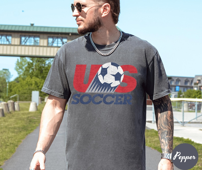 May include: A charcoal gray t-shirt with the text "US Soccer" in red and blue, featuring a soccer ball graphic. The shirt has a vintage, worn look.